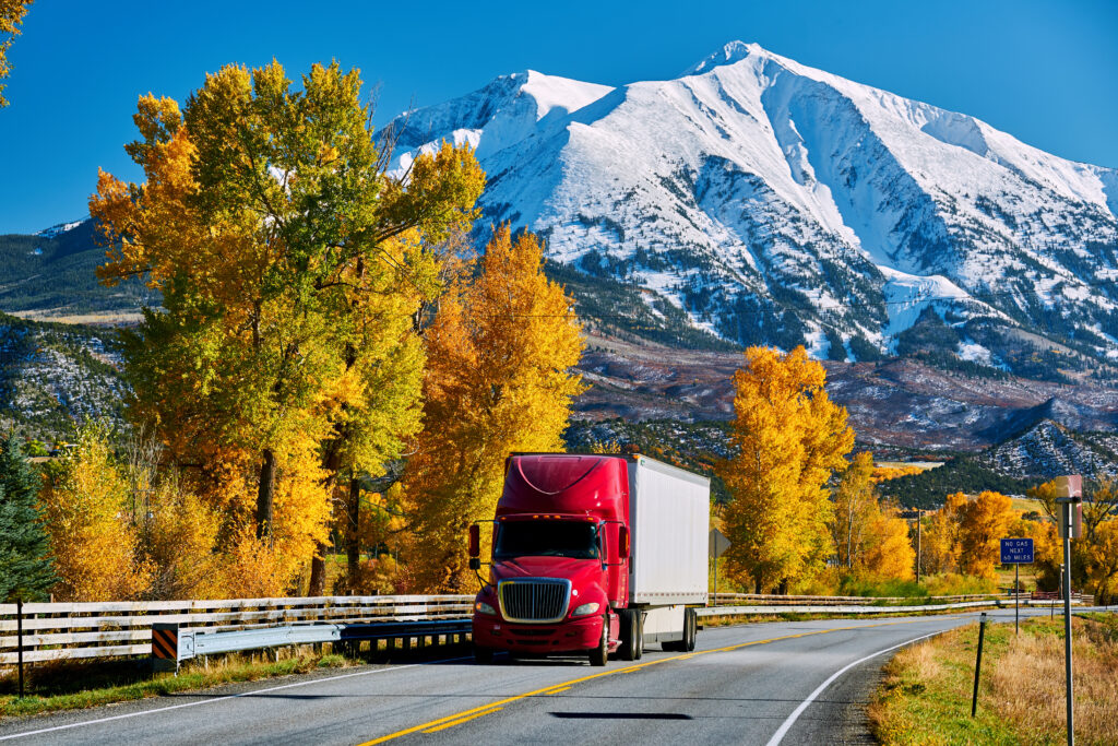 Semi truck driving on country road with colorful fall leaves on a nearby tree and snow capped mountains in the background